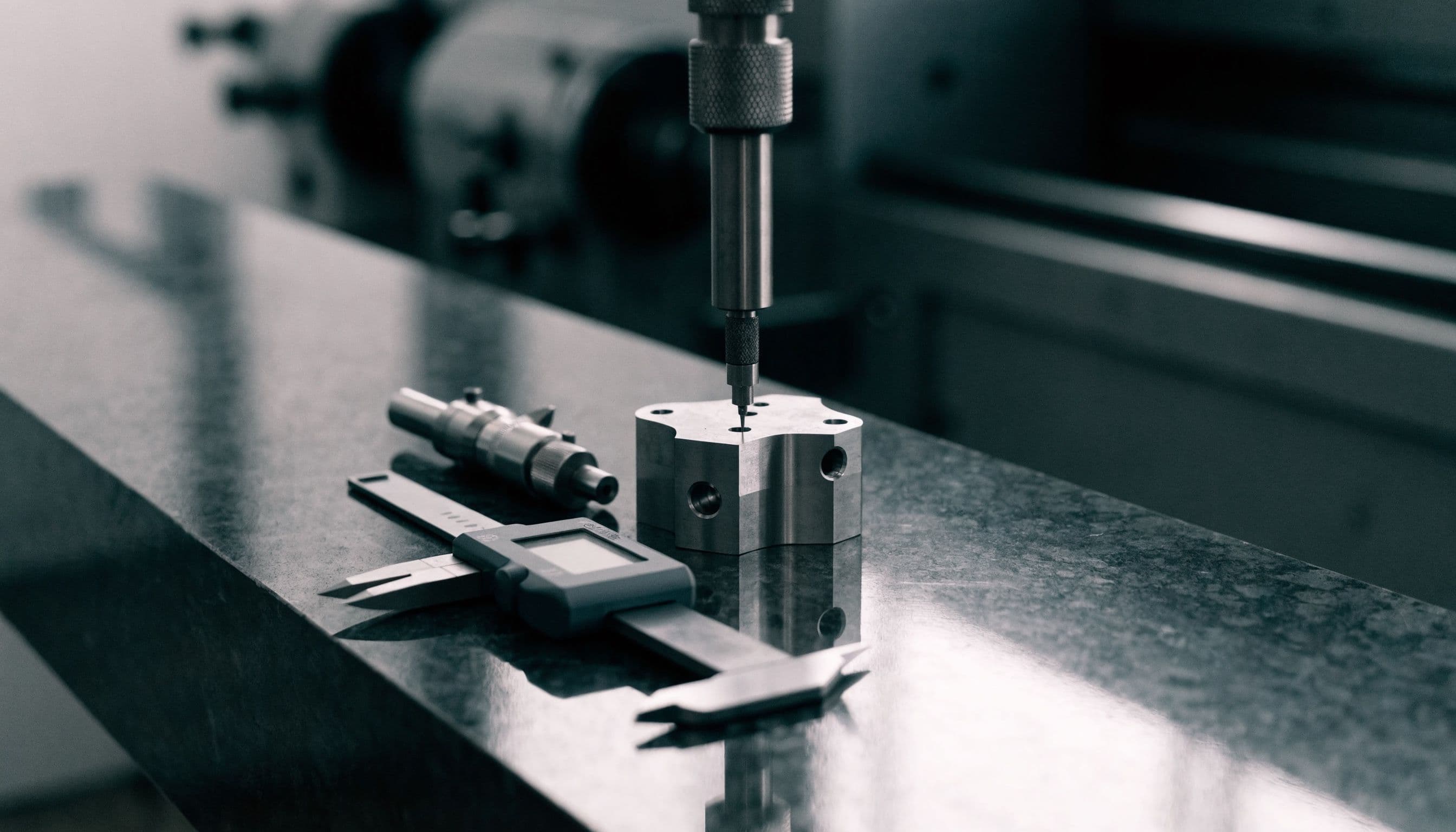 A machined metal part on a granite surface plate being measured with a precision height gauge, beside digital calipers and a micrometer.